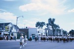 A band is marching on Newton's Main Street during the 1962 Harvey County Fair parade with people watching from the side of the street; a Valentine diner (R. & T. Lunch) is seen in the far left background.