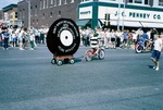 A boy riding a bicycle is pulling a wagon that has a cardboard cutout that looks like a tire during the 1965 Harvey County Fair parade on Main Street in Newton