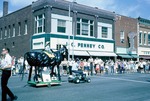A boy is sitting on a replica of a horse made from tires which is being pulled by a small garden tractor down Newton's Main Street during the 1965 Harvey County Fair parade; a crowd of people are watching from the side of the street