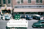 4-H float in the 1954 Harvey County Fair parade being pulled by a car in the 600 block of North Main in Newton; several children are sitting in the float and there are people and automobiles along the side of the street