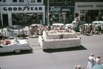 4-H float being pulled by a convertible in the 1956 Harvey County Fair parade; people and automobiles are in front of some of the downtown businesses: Goodyear, Reese Drugs and Marietta Shop