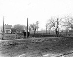 A bull is standing inside a fenced area with a frame house directly behind the fenced area. Railway tracks, grain elevator, and a few other business buildings are visible in the distance